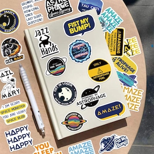 May include: A light-coloured notebook covered in various space-themed stickers, including text like FIST MY BUMP, JAZZ HANDS, and AMAZE, alongside a white pen. The stickers feature colourful planets, astronauts, and warning signs with bold typography.