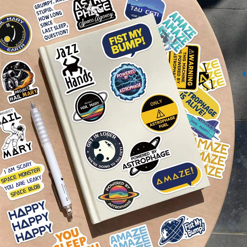 May include: A light-coloured notebook covered in various space-themed stickers, including text like FIST MY BUMP, JAZZ HANDS, and AMAZE, alongside a white pen. The stickers feature colourful planets, astronauts, and warning signs with bold typography.