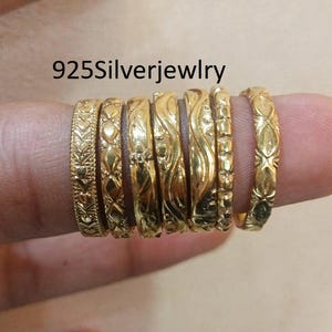 Puede incluir: Un conjunto de siete anillos de tono dorado con varios patrones en relieve, incluyendo diseños de corazones, geométricos y ondulados. Los anillos están dispuestos en fila y el texto 925Silverjewlry es visible arriba. Cada anillo tiene textura.