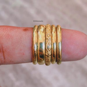 May include: A set of five gold-toned rings arranged in a row, featuring four smooth bands and one central band with an intricate embossed floral pattern. The text 925SilverJewelry is visible above the rings, which are displayed on a finger.