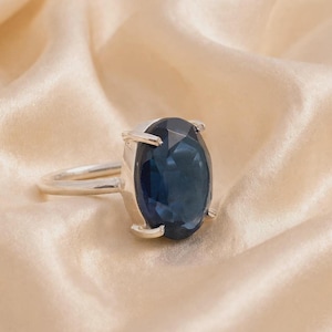 May include: A silver ring featuring a large, oval-cut, deep blue gemstone held in place by four prongs. The band is smooth and polished, showcasing a simple design that highlights the dark blue stone as the central focal point of the piece.