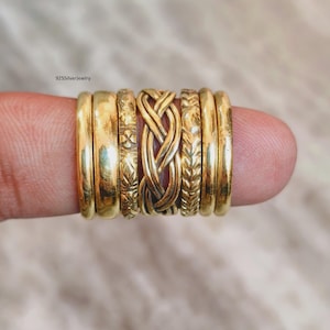 May include: A set of seven gold-toned rings featuring various designs, including smooth bands, a braided centre ring, and bands with floral and leaf-like patterns. The text 925silverjewelry is visible in the upper left corner of the image.