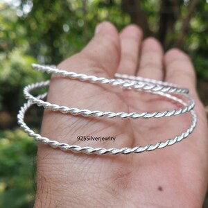 Puede incluir: Una pulsera tipo brazalete de plata con un diseño de cuerda trenzada. La superficie metálica es pulida y reflectante. El texto 925Silverjewelry está impreso en negro sobre la superficie de la pulsera, indicando la composición del material.