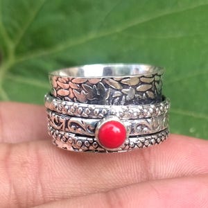 May include: A silver spinner ring featuring a wide band with an oxidised floral pattern, a central rotating band with a textured dotted finish, and a smaller band adorned with a single round, smooth, bright red coral gemstone set in silver.