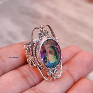 Puede incluir: Un anillo de plata de ley con una piedra preciosa de topacio místico iridiscente de corte ovalado engastada en un bisel de cuerda trenzada. La banda y el engaste muestran detalles metálicos intrincados de filigrana. El texto 925Silverjewelry es visible.