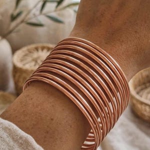 Puede incluir: Un conjunto de múltiples brazaletes circulares y delgados de color cobre que se usan juntos en la muñeca. Las pulseras presentan un acabado metálico liso y pulido con un diseño simple de alambre redondeado que crea un aspecto apilado.