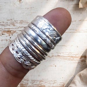Könnte beinhalten: Ein Stapel aus sechs Silberringen an einem Finger. Der oberste Ring hat ein geprägtes Blumenmuster, gefolgt von drei glatten, polierten Bändern, einem gedrehten Seilband und einem unteren Ring mit einem komplexen Flechtmuster.