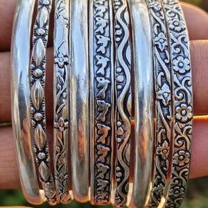 Puede incluir: Un conjunto de ocho brazaletes de plata con varios diseños, incluyendo patrones florales, líneas onduladas y motivos de aves repetidos. El texto 925Silverjewelry es visible en la parte superior. Cada brazalete tiene un acabado metálico pulido con detalles oxidados.