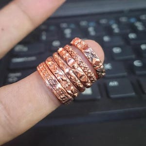 May include: A stack of six copper-toned metal rings worn on a finger. Each ring features a distinct embossed pattern, including geometric shapes, floral motifs, and heart designs, with a polished finish that reflects light across the textured surfaces.