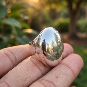 Puede incluir: Un anillo de tono plateado pulido que presenta una cúpula grande, lisa y de forma ovalada que actúa como superficie reflectante. La banda de metal es sólida y continua, con la parte superior redondeada creando un punto focal convexo.