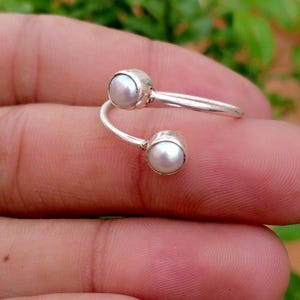 May include: A silver bypass ring featuring two small, round, white pearls set at each end of the open band. The polished silver metal band curves around to hold the pearls in place, creating a simple, open-ended design that is adjustable in size.