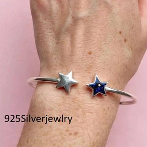 May include: A silver cuff bracelet featuring two star-shaped ends. One star is solid polished silver, while the other star is inlaid with a dark blue speckled stone. The text 925Silverjewelry is printed in black in the lower left corner of the image.