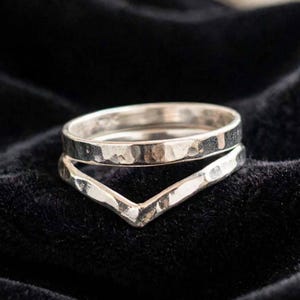 Könnte beinhalten: Ein Set aus zwei silbernen Ringen mit gehämmertem Muster. Ein Ring ist ein einfacher kreisförmiger Bandring, während der andere eine V-förmige Chevron-Form aufweist. Beide Ringe haben eine polierte Metalloberfläche und eine unebene Struktur.