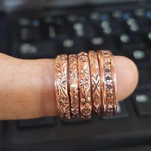 May include: A set of five copper-colored rings featuring various embossed patterns, including floral motifs, geometric shapes, and a repeating cross design. Each ring has a distinct texture and metallic finish, displayed together in a row.