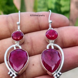 May include: A pair of sterling silver drop earrings featuring faceted, deep pinkish-red gemstones. Each earring has a small round stone above a larger pear-shaped stone, both set in silver frames with curved accents and the text 925Silverjewlry.