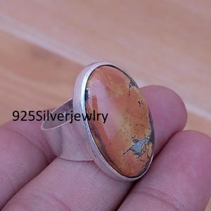 May include: A sterling silver ring featuring a large, oval-shaped, polished orange and brown stone with dark veining. The band is wide and smooth. The text 925Silverjewelry is visible on the side of the ring, indicating the metal composition.