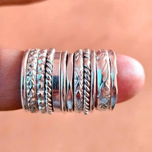 May include: A collection of ten silver rings featuring various textures and patterns, including braided, beaded, smooth, and engraved designs. The rings are arranged side-by-side, showcasing a range of metallic finishes and intricate details.