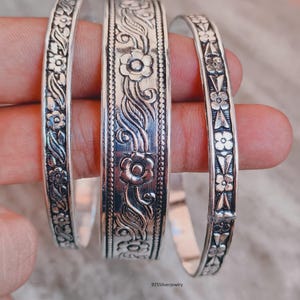 Puede incluir: Tres brazaletes de plata con intrincados patrones florales y de enredaderas en relieve. El brazalete central es el más ancho, mientras que los dos laterales son más estrechos. Cada pieza tiene un acabado pulido con detalles oscuros.
