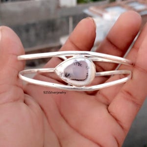 May include: A silver bracelet with a teardrop-shaped, white and gray stone. The stone is set in a silver bezel and is the focal point of the bracelet. The bracelet has a simple, elegant design.