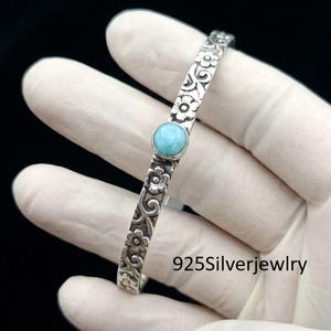 May include: A silver cuff bracelet featuring an embossed floral pattern and a single round, light blue stone set in the centre. The metal surface has darkened recessed areas to highlight the flower details. The text 925Silverjewlry is visible.