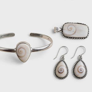 Op de afbeelding: Een sieradenset bestaande uit een zilveren armband, een rechthoekige hanger en een paar druppelvormige oorbellen. Elk sieraad bevat een witte shiva-oogschelp met een bruin spiraalpatroon, gezet in 925 sterling zilver met gedraaide randen.