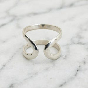 May include: A polished silver-toned ring featuring an open, bypass-style design. The band consists of two smooth, curved ends that overlap slightly without touching, creating a continuous, fluid shape with a reflective, metallic surface finish.