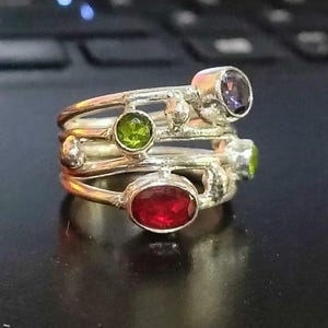 May include: A multi-band silver ring featuring a textured, organic metal finish. The ring is set with four distinct gemstones: a large oval red stone, a round green stone, a smaller round green stone, and a round purple stone at the top.
