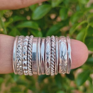 May include: A collection of eleven silver rings stacked together, featuring various textures including smooth bands, twisted rope patterns, engraved floral motifs, geometric cross-hatch designs, and beaded details, all crafted from polished silver metal.