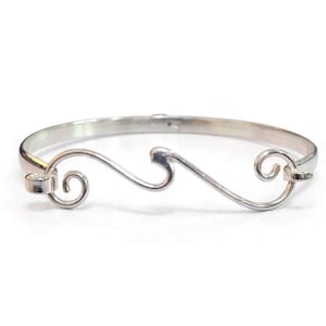 May include: A polished silver-tone bangle bracelet featuring a central wave-like design with two spiraling ends. The metal band is smooth and circular, with the decorative wave element integrated into the front section of the jewelry piece.