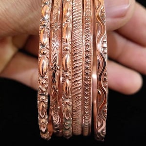 May include: A set of five copper-colored bangles featuring distinct embossed patterns. The bracelets display varied textures including repeating oval shapes, cross-hatch designs, and wavy lines, all finished with a polished metallic sheen.