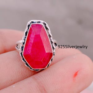 Può includere: Un anello in argento con una grande pietra preziosa rossa sfaccettata a forma di bara incastonata in una fascia di metallo intrecciato. L'anello riporta la scritta 925Silverjewelry. La pietra ha una tonalità rosso rosato intensa e vivace.