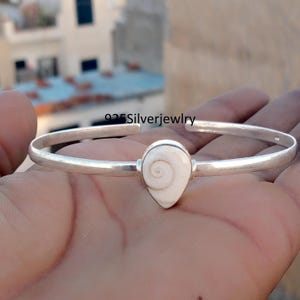 Può includere: Un bracciale in argento con un elemento centrale a forma di goccia di conchiglia bianca. Il bracciale è un semplice design a polsino aperto. Il testo "925 Silverjewlry" è visibile.