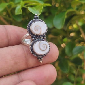 Peut inclure: Une bague en argent ornée de deux coquillages Shiva eye ovales superposés, chacun serti dans une bordure en fil d'argent torsadé. L'anneau est divisé et le sertissage comprend de petites perles d'argent aux extrémités supérieure et inférieure.
