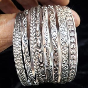 May include: A set of seven silver-toned metal bangles featuring various embossed patterns including geometric dots, wavy lines, leaf-like textures, floral motifs, and a repeating heart design, all with an oxidized finish for depth.
