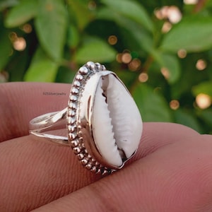 May include: A silver ring featuring a white cowrie shell centerpiece set within a beaded metal border. The band is split into two thin silver segments. The text 925Silverjewelry is visible on the side of the ring, indicating the metal composition.