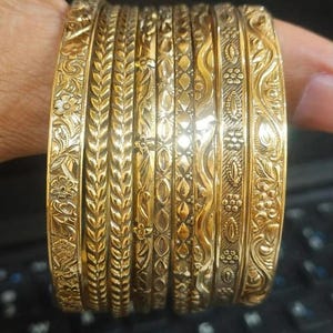 Op de afbeelding: Een set van acht goudkleurige metalen armbanden met diverse reliëfpatronen, waaronder bloemmotieven, bladachtige texturen, geometrische ruitvormen en golvende lijnen. Elke armband heeft een eigen ontwerp met een gepolijste afwerking.