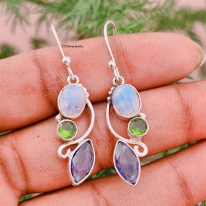 Puede incluir: Un par de pendientes colgantes de plata de ley con una disposición vertical de tres piedras preciosas: una piedra lunar redonda iridiscente, un pequeño peridoto verde redondo y una amatista morada en talla marquesa.