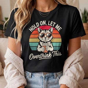 Op de afbeelding: Zwart t-shirt met een kat met een hartvormige zonnebril, die een drankje vasthoudt, en de tekst "HOLD ON, LET ME Overthink This" tegen een retro zonsondergang achtergrond.
