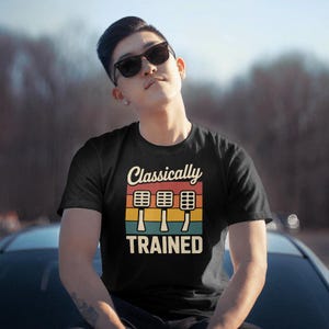 Może przedstawiać: Czarny t-shirt z napisem "Classically TRAINED" i grafiką trzech pedałów samochodowych w kolorze czerwonym, żółtym i niebieskim. Koszulka jest noszona przez osobę w okularach przeciwsłonecznych.