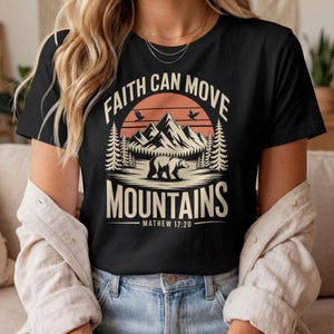 Peut inclure: T-shirt noir avec un motif graphique représentant une chaîne de montagnes, un ours et le texte "FAITH CAN MOVE MOUNTAINS - MATHEW 17:20". Le motif comprend un coucher de soleil orange et deux oiseaux. Le t-shirt est à col rond.