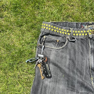 Peut inclure: Jean en denim gris foncé avec une ceinture à motif d'étoiles jaunes, une attache de passant de ceinture en forme de cœur et un mousqueton en métal argenté retenant un trousseau de clés, dont une clé ancienne en laiton et une clé de voiture noire.