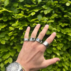 May include: A hand displaying five silver rings of different styles. The hand also has a silver watch and a green bracelet. The backdrop is a wall of green foliage.