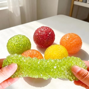 May include: A collection of five textured, translucent, spherical objects in red, orange, yellow, and two shades of green. One elongated, flexible green piece is held in the foreground, while four round spheres are arranged behind it on a surface.