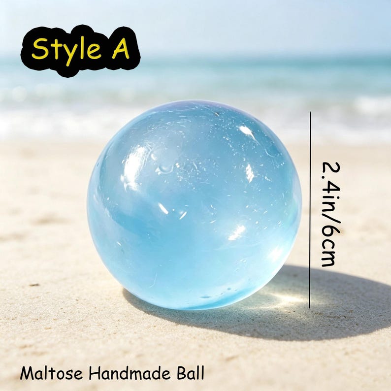 May include: A translucent light blue spherical object labeled Style A and Maltose Handmade Ball. The sphere has a smooth, glossy surface and a diameter of 2.4 inches. The object is shown against a bright, out-of-focus coastal background.