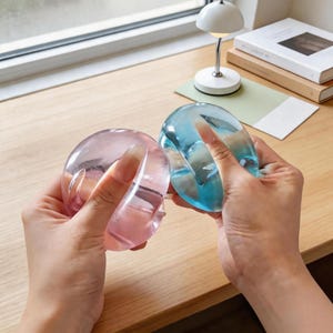 May include: Two clear, rounded bangles, one pink and one blue, are held up against each other. The bangles have a smooth, glossy finish. The background includes a desk, a small lamp, and books.