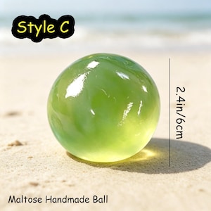 May include: A translucent, light green, spherical Maltose handmade ball labeled Style C. The object has a smooth, glossy surface with visible light reflections. A vertical line indicates a diameter measurement of 2.4 inches.