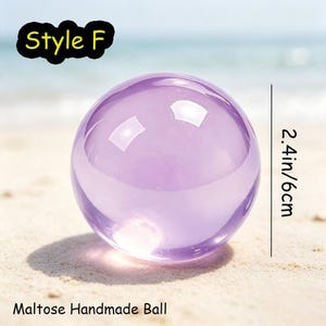 May include: A smooth, translucent purple sphere labeled Style F and Maltose Handmade Ball. The object is 2.4 inches in diameter, featuring a clear, glass-like appearance with light reflections on its curved surface, set against a bright background.