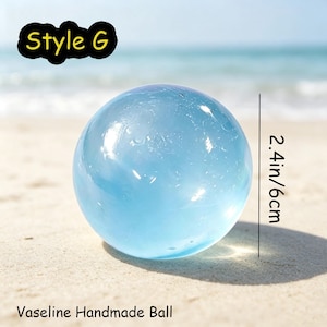 May include: A light blue, translucent, spherical handmade ball labeled Style G. The ball has a diameter of 2.4 inches. The text Vaseline Handmade Ball is printed at the bottom of the image, which shows the object against a beach background.