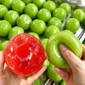 May include: A glossy, translucent red sphere with small black specks and a glossy, translucent green ring-shaped object with small black specks, both held against a background of numerous small, round, bright green spheres.