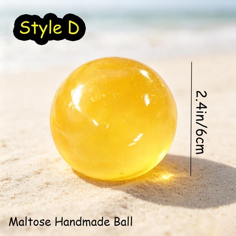May include: A translucent yellow spherical Maltose handmade ball labeled Style D. The object has a smooth, glossy surface and a diameter of 2.4 inches as indicated by the vertical measurement line next to the sphere.